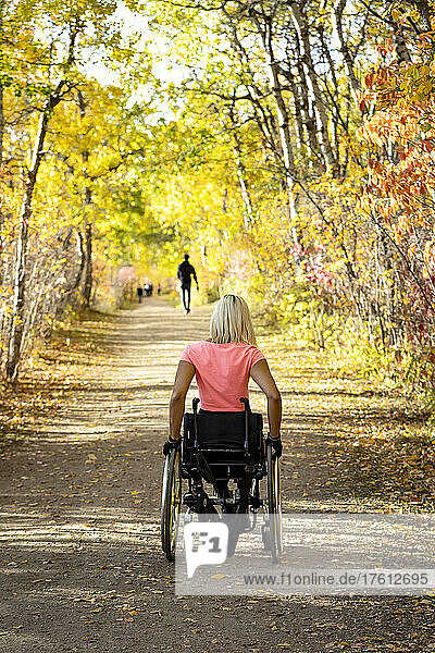 Young paraplegic woman in her wheelchair going down a trail in a park on a beautiful fall day; Edmonton  Alberta  Canada