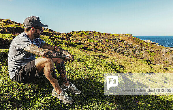 Mann mit Tattoos  Hut und Sonnenbrille sitzt auf einem grasbewachsenen Hügel am Nakalele Point mit Blick auf den Ozean; Maui  Hawaii  Vereinigte Staaten von Amerika