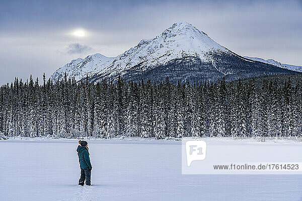 Frau  die im Schnee steht und die Aussicht betrachtet  während sie auf dem zugefrorenen Tagish Lake spazieren geht  während die Sonne durch die Wolken über den Berggipfeln lugt; Tagish  Yukon  Kanada