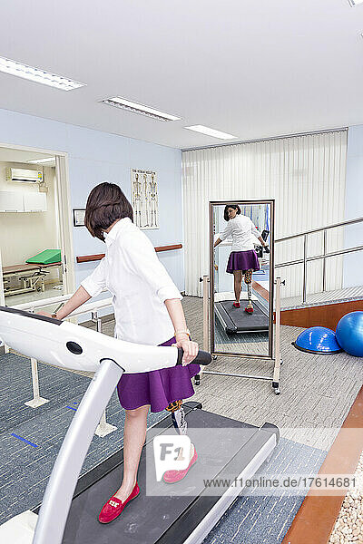 Young woman with leg prosthesis in therapy  walking on a treadmill and looking in the mirror at her reflection as she walks; Bangkok  Thailand