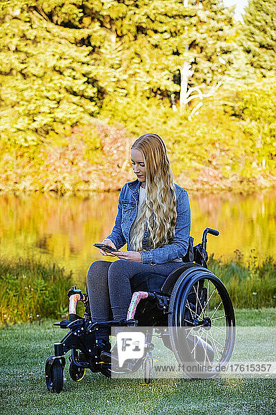Junge querschnittsgelähmte Frau in ihrem Rollstuhl benutzt ein Smartphone in einem Park an einem schönen Herbsttag; Edmonton  Alberta  Kanada