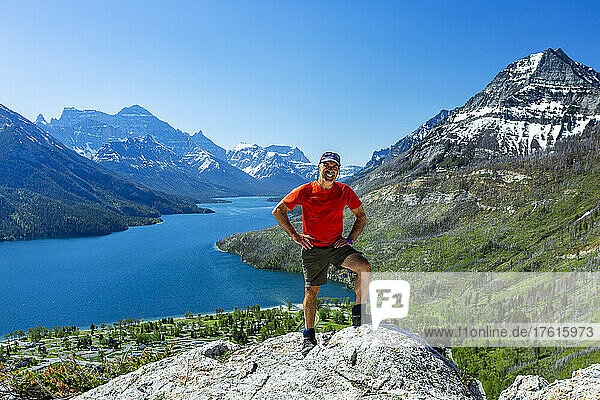 A male hiker standing on top of a rock cliff with mountains and a lake in the distance with blue sky  Waterton Lakes National Park; Waterton  Alberta  Canada