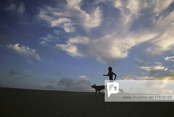 A woman and her dog run along the beach with beautiful sky.; Jockeys Ridge State Park  Nags Head  North Carolina