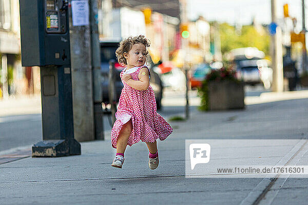 Preschooler girl running down a city sidewalk in her red and white gingham dress; Toronto  Ontario  Canada