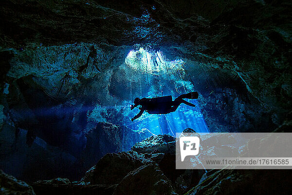 A cave diver descends into a cave system with light rays penetrating the water in the background.
