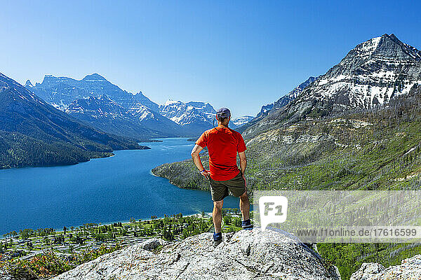 A male hiker standing on top of a rock cliff overlooking the mountains and a lake in the distance with blue sky  Waterton Lakes National Park; Waterton  Alberta  Canada