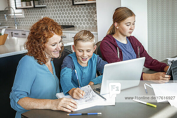 A teenage girl and young boy sit at the kitchen table at home with a laptop and tablet doing school work while being homeschooled; Alberta  Canada