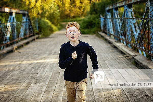 Young boy with red hair on a park bridge runs towards the camera; Edmonton  Alberta  Canada