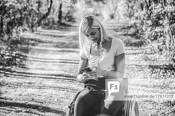 Junge querschnittsgelähmte Frau in ihrem Rollstuhl auf einem Weg in einem Park  die ihr Smartphone benutzt; Edmonton  Alberta  Kanada
