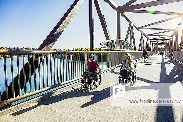 Young paraplegic man and woman going across a bridge together using their wheelchairs on a beautiful fall day; Edmonton  Alberta  Canada