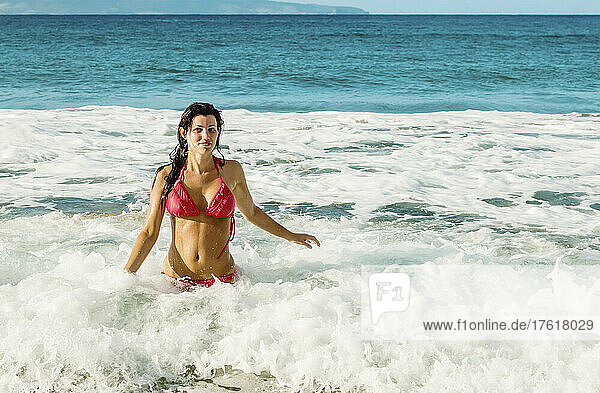 Frau im roten Bikini steht im Meerwasser vor der Küste und schaut in die Kamera am D. T. Fleming Beach mit der Insel Molokai in der Ferne; Kapalua  Maui  Hawaii  Vereinigte Staaten von Amerika