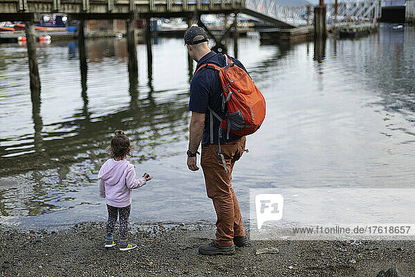 Father and young daughter stand at the water's edge of a beach looking out at the harbour of Horseshoe Bay in BC  Canada; West Vancouver  British Columbia  Canada