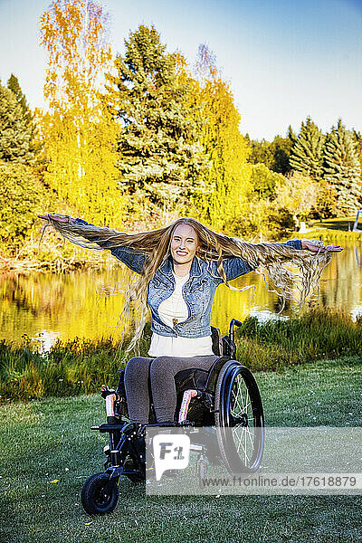 Junge querschnittsgelähmte Frau in ihrem Rollstuhl in einem Park an einem schönen Herbsttag. Sie fühlt sich unbeschwert  streckt ihre Arme aus und zeigt die Länge ihrer langen Haare; Edmonton  Alberta  Kanada