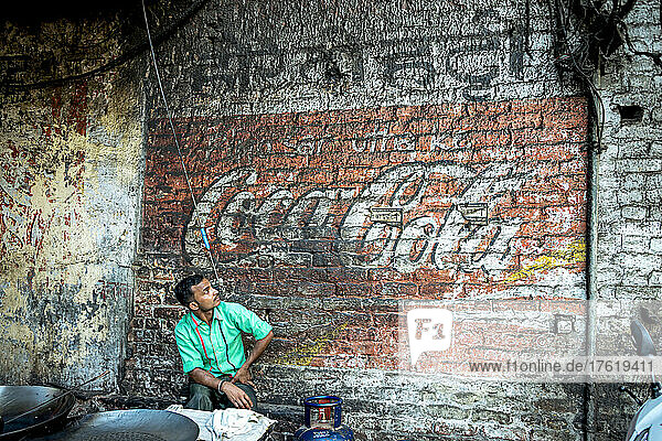 Mann sitzt und schaut auf ein verblasstes Cola-Schild  das auf eine Backsteinmauer gemalt ist; Amritsar  Punjab  Indien