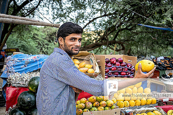 Junger Mann  der frisches Obst in die Kamera hält  an einem Obststand  der frische Produkte verkauft; Greater Noida  Uttar Pradesh  Indien