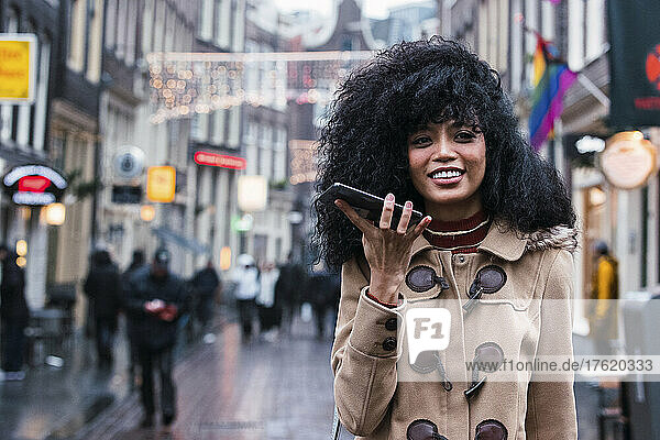Happy woman with tousled hair talking on mobile phone through speaker in city
