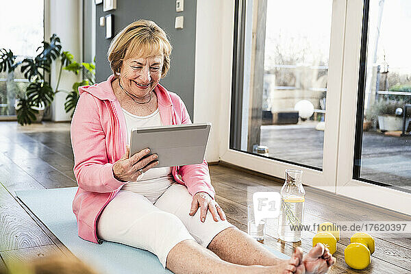Smiling blond senior woman with tablet PC sitting on exercise mat at home