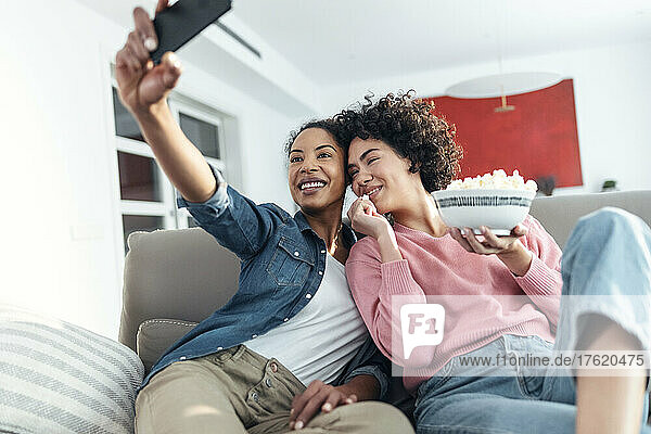Frau macht ein Selfie mit einer Freundin  die zu Hause auf dem Sofa Popcorn isst