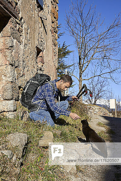 Man with backpack playing with street cat on sunny day