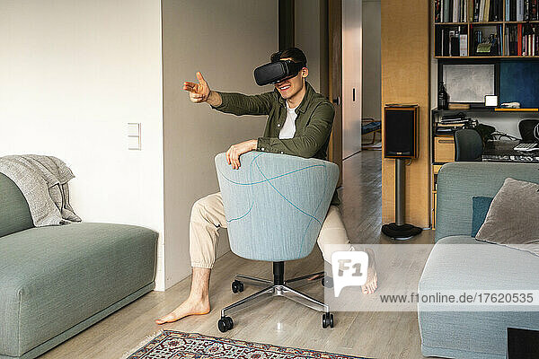 Smiling young man wearing virtual reality simulator gesturing on chair at home