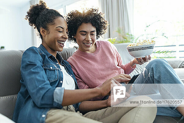 Happy woman sharing smart phone with friend sitting on sofa in living room