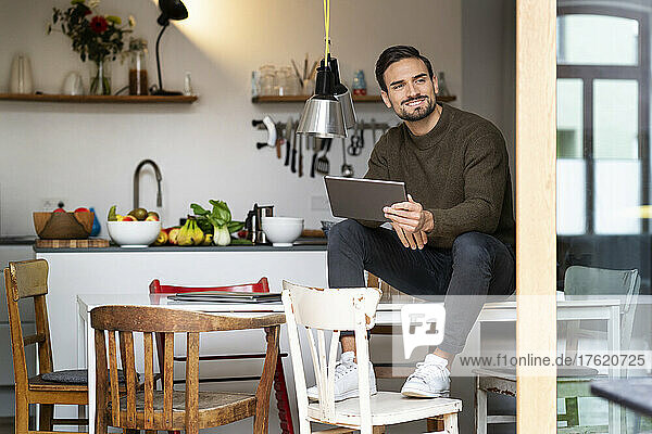 Lächelnder junger Mann mit Tablet-PC sitzt zu Hause am Tisch in der Küche