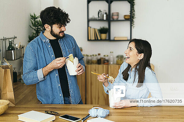 Smiling woman and man eating noodles with chopsticks in kitchen at home