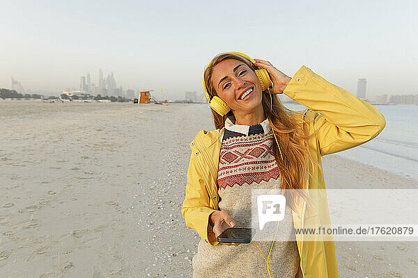 Glückliche Frau in gelber Jacke  die am Strand Musik über kabellose Kopfhörer hört