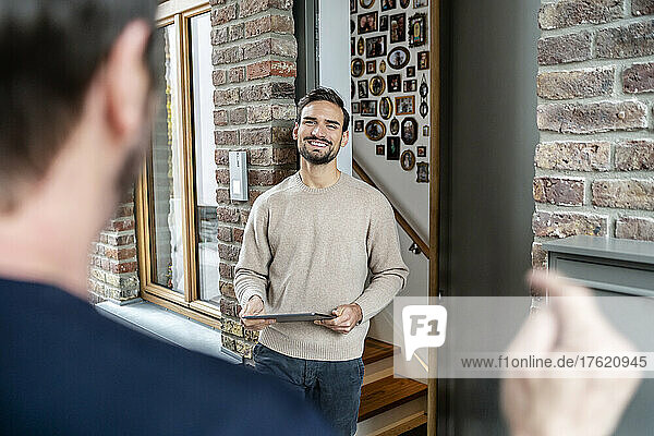 Smiling man holding tablet PC talking with friend outside house