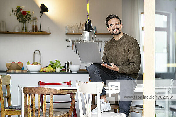 Smiling freelancer with laptop sitting on table in kitchen at home