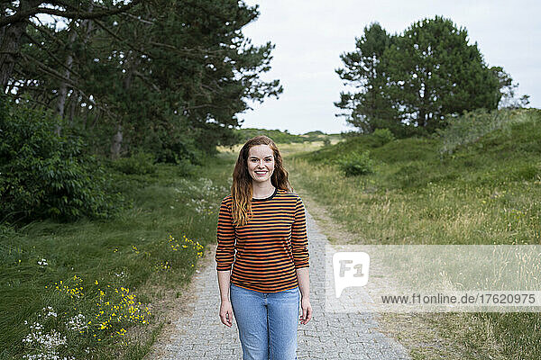 Happy redhead woman standing on footpath amidst meadow