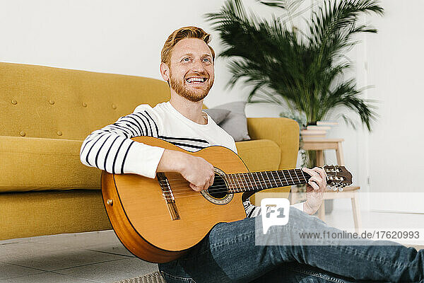 Fröhlicher Mann mit Gitarre sitzt zu Hause vor dem Sofa