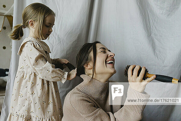 Fröhliche Frau mit Haarbürste singt Tochter  die zu Hause Haare bindet