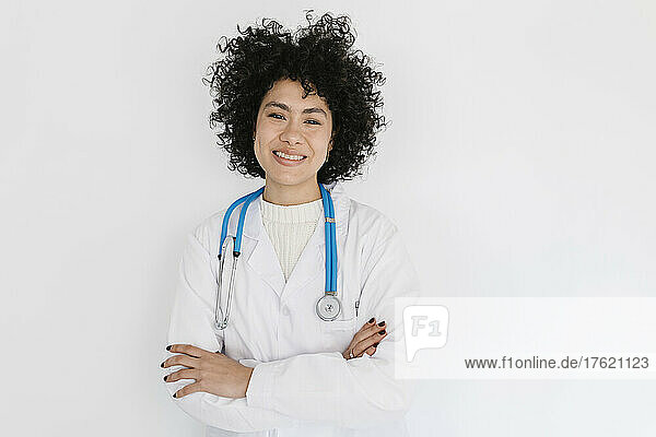 Smiling doctor standing with arms crossed in front of white wall