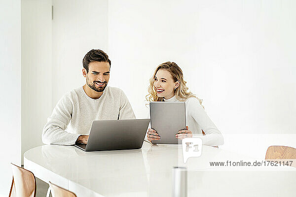 Smiling blond woman with tablet PC looking at colleague using laptop