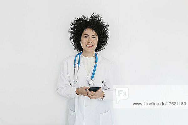 Smiling doctor holding mobile phone standing in front of white wall