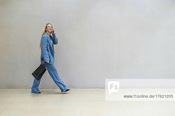 Smiling businesswoman with shopping bag talking on smart phone walking by wall