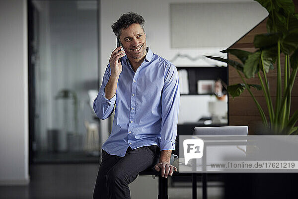 Happy businessman talking on smart phone sitting on desk in office