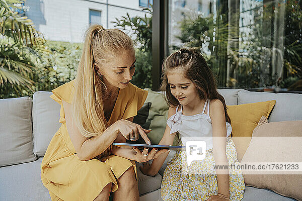 Mother sharing tablet PC with daughter sitting on sofa at patio