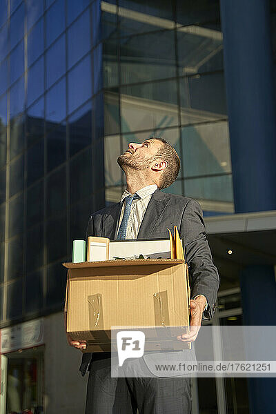 Businessman with box looking up by office building on sunny day