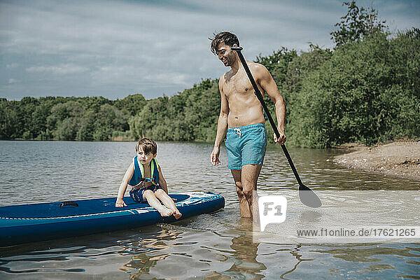Son sitting on paddleboard and father standing in lake