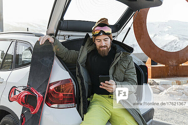 Young man with snowboard sitting in car trunk using smart phone
