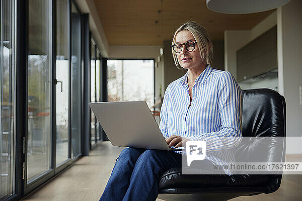 Blonde Frau mit Brille und Laptop sitzt zu Hause auf einem Stuhl
