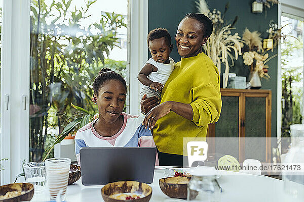 Smiling mother looking at daughter studying on tablet PC in dining room