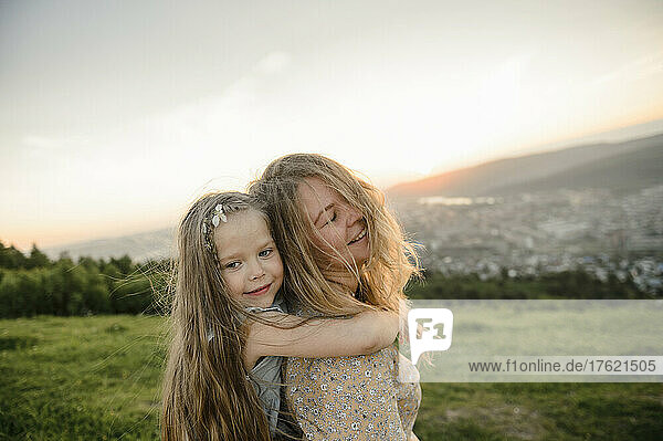 Süßes Mädchen mit blonden Haaren umarmt Mutter bei Sonnenuntergang