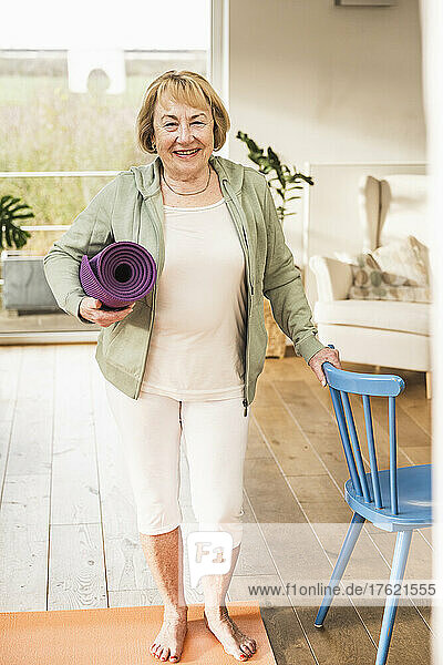 Happy senior with exercise mat standing by chair at home