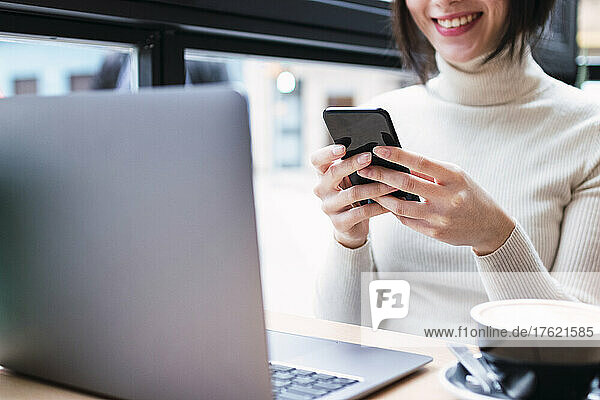 Lächelnde Geschäftsfrau mit Laptop und Mobiltelefon im Café sitzend