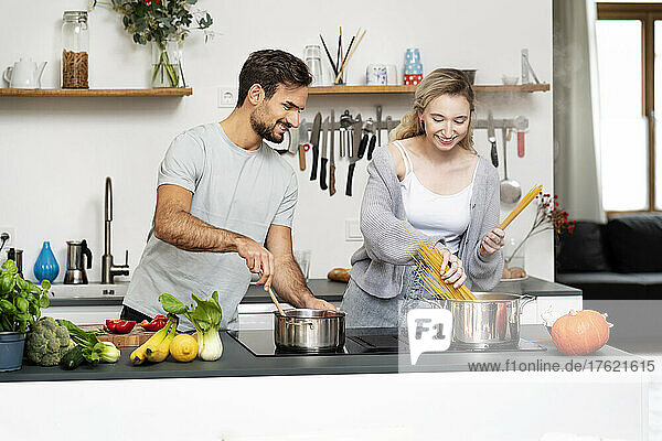 Smiling young man looking at girlfriend making pasta in kitchen