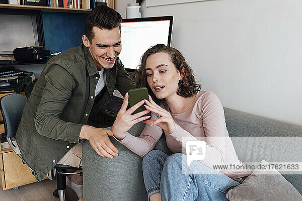 Junge Frau teilt Smartphone mit Freund  der auf einem Stuhl im Wohnzimmer sitzt