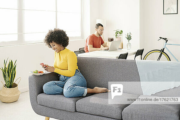 Lächelnde junge Frau benutzt Smartphone  während Mann mit Laptop im Wohnzimmer sitzt